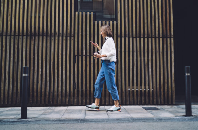 Focused woman strolling on sidewalk and reading message on smartphone